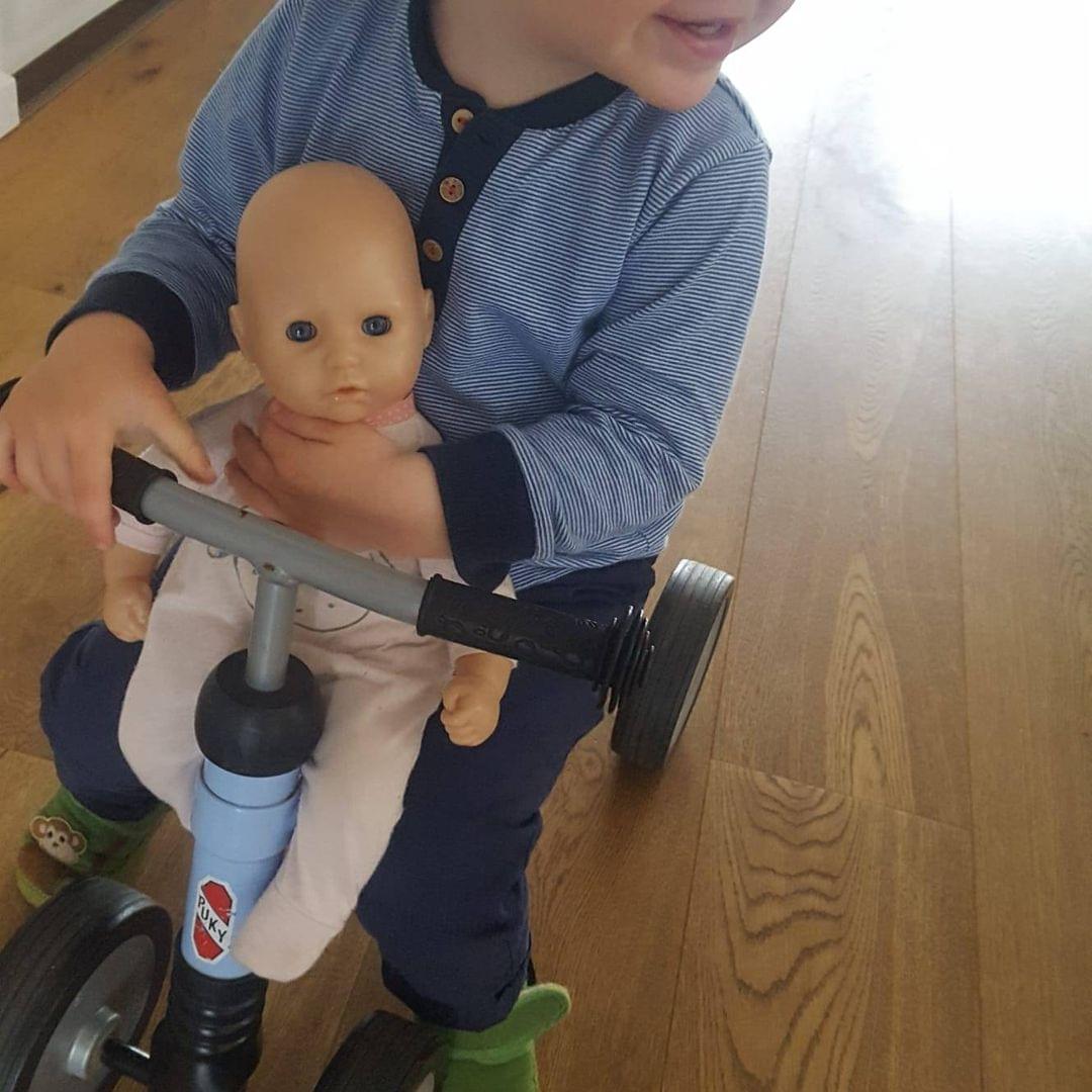 Foto eines Kindes, das mit einer Puppe ein Rutschfahrzeug fährt